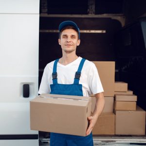 Deliveryman with carton box at the car, delivery service. Man in uniform holding cardboard package, male deliver, courier job
